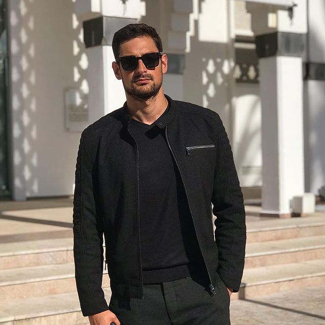 Brice Bexter in a black t-shirt, outer, and black pants posing in front of his house.